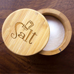 The "Salt Shaker" Engraved Salt Cellar by Totally Bamboo, with its swiveling lid featuring a salt shaker icon and magnetic lock, is open to display white salt inside against a dark wooden surface.