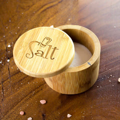 A "Salt Shaker" Engraved Salt Cellar by Totally Bamboo, boasting a 6-ounce capacity and designed with a swiveling lid, slightly open to reveal fine white salt inside. The word "Salt" is engraved on the lid. This container features a magnetic lock and rests on a wooden surface with a few scattered grains of salt around it.