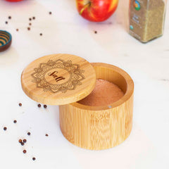 A round salt box crafted from wood by Totally Bamboo can be seen with its magnetic lid slightly open, showing pink salt inside. The engraved lid features a decorative mandala pattern with "Salt" at its center. In the background, various spices and an apple appear blurred, enhancing the elegance of this 6-ounce capacity "Mandala Salt" Engraved Salt Cellar.