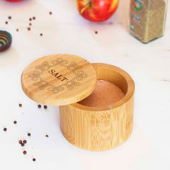 A "Celtic Knot" Engraved Salt Cellar from Totally Bamboo, partially open to showcase pink Himalayan salt inside its secure magnetic-closure lid, sits on a white surface. The scene is completed by whole black peppercorns, a red apple, and a container of spices surrounding it.