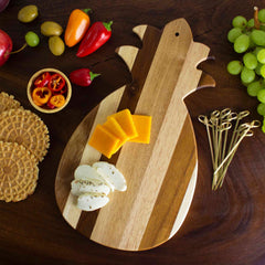 The Rock & Branch® Shiplap Series Pineapple Shaped Cutting Board by Totally Bamboo displays slices of cheddar and mozzarella cheese. It's encircled by vibrant fruits, waffles, sliced peppers, and skewers on a dark wooden surface.