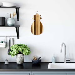 A contemporary kitchen features light gray cabinets, a black countertop, and a silver faucet. A potted green plant and wooden bowl contribute to the Tropical Style ambiance. Shelves display dishes and utensils, while a Totally Bamboo Rock & Branch® Shiplap Series Pineapple Shaped Cutting Board adorns the white wall.