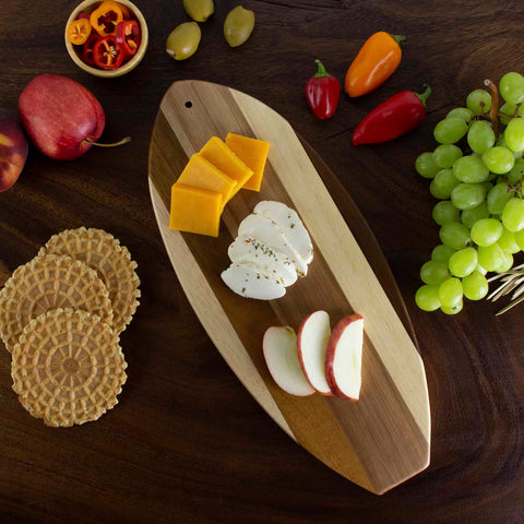 The Rock & Branch® Shiplap Series Li'l Surfer Cutting Board from Totally Bamboo provides a charming rustic display, featuring an assortment of white and cheddar cheese slices, an apple, green grapes, vibrant red and yellow peppers, a bowl of sliced peppers, and waffle cookies arranged on a wooden table. It's perfect for bringing charm to your kitchen or serving as delightful wall decor!