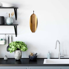 A modern kitchen featuring a dark countertop hosts a sleek sink with a metallic faucet. Shelves are lined with dishes and kitchenware, while the Rock & Branch® Shiplap Series Li'l Surfer Cutting Board, shaped like a surfboard from Totally Bamboo, hangs on the wall adding flair to the display of wall art. Beside a mortar and pestle on the counter sits a potted plant.