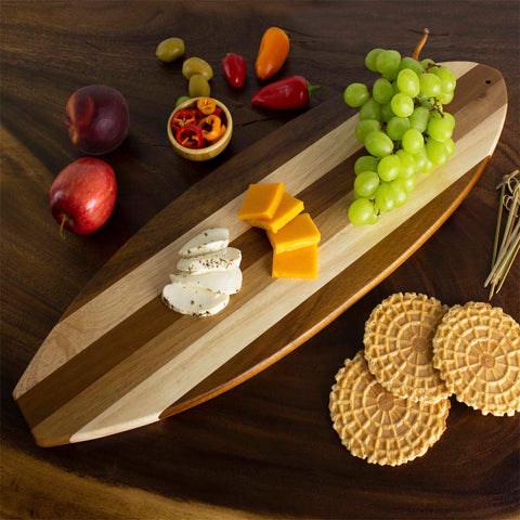 A Rock & Branch® Shiplap Series Surfboard Shaped Cutting Board by Totally Bamboo, teeming with grapes, cheddar, and herbed cheese, is encircled by crisp wafers, plums, olives, and mini peppers on a dark wooden table.