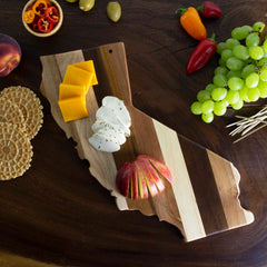 A Rock & Branch® Shiplap Series California Cutting Board by Totally Bamboo elegantly displays slices of cheddar and mozzarella cheese and a decorative apple. The wooden board is surrounded by grapes, crackers, and assorted peppers, crafting a delightful edible display that doubles as perfect wall art when not in use.