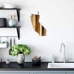 A kitchen interior with a modern aesthetic features the Rock & Branch® Shiplap Series California Cutting Board by Totally Bamboo, which doubles as wall art. Below, Rock & Branch Shiplap shelves hold kitchen items, while the countertop boasts a potted plant and a sleek faucet by the sink.