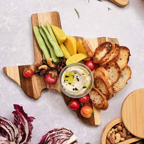 The Rock & Branch® Shiplap Series Texas Cutting Board by Totally Bamboo features vibrant wood plank colors and is elegantly topped with sliced cucumbers, yellow bell peppers, radishes, tomatoes, crostini, and nuts. It is accompanied by a glass bowl of dip and surrounded by radicchio leaves along with a small bowl of nuts.