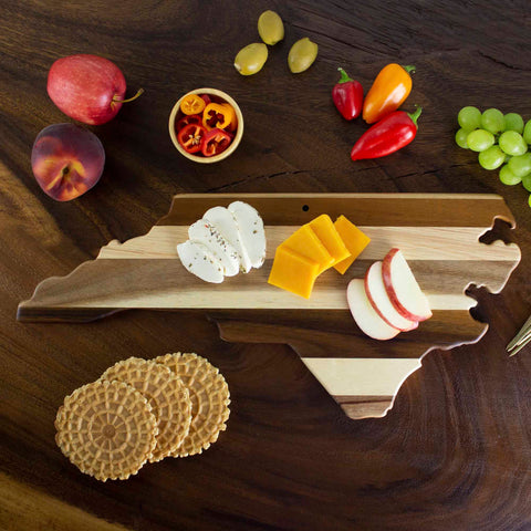 A Rock & Branch® Shiplap Series North Carolina Cutting Board by Totally Bamboo rests on a rustic wood plank, featuring sliced cheese, apples, and crackers. Nearby, vibrant red and orange peppers, grapes, apples, and a bowl of sliced peppers complete the delightful spread that beautifully complements the wall art.