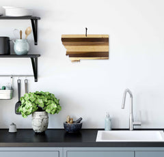 Modern kitchen featuring a minimalist design with a sleek black countertop and faucet. The Rock & Branch® Shiplap Series Montana Cutting Board by Totally Bamboo hangs as wall art, shaped like the state of Montana. Shelves hold dishes and a potted plant, adding greenery, while the Shiplap Series adds texture to the decor.