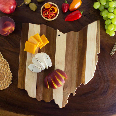 The Rock & Branch® Shiplap Series Ohio Cutting Board from Totally Bamboo, shaped like the state of Ohio, is adorned with cheese slices and an apple. Grapes, peppers, a nectarine, and a small bowl of sliced peppers are arranged around it on a dark table that highlights its unique shiplap design.