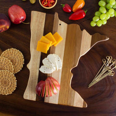 A Rock & Branch® Shiplap Series Minnesota Cutting Board by Totally Bamboo holds an assortment of cheeses, sliced apples, and crackers. Grapes, peppers, cherry tomatoes, and small biscuits are artfully spread across a rustic wood plank table surrounding the cutting board.