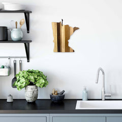 Modern kitchen featuring a black countertop, gray cabinets, and a white sink. A plant and decorative items rest on the counter. Wooden shelves display kitchenware, while the Rock & Branch® Shiplap Series Minnesota Cutting Board from Totally Bamboo adds charm to the decor in its unique U.S. state shape.