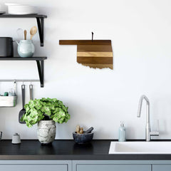 A kitchen showcases a black countertop with a sink, enhanced by a plant in a stone vase and a small bowl. Above, the Shiplap Series shelf displays kitchen items, while the white wall features the Rock & Branch® Shiplap Series Oklahoma Cutting Board by Totally Bamboo, shaped like the outline of Oklahoma.