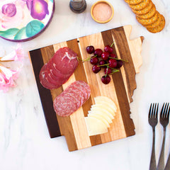 A Rock & Branch® Shiplap Series Arkansas Cutting Board by Totally Bamboo holds sliced meats, cheese, and cherries. Nearby are crackers, forks, a small bowl of salt, lilies, and a crockery dish with a floral design set on a marble surface. The board’s wood plank colors evoke the charm of Rock & Branch Shiplap wall art.