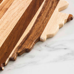 On a white marble surface lies a close-up of the Rock & Branch® Shiplap Series Arkansas Cutting Board by Totally Bamboo. This multicolored wooden board, with its distinctive wavy edge, features alternating strips of light and dark wood that reveal a stunning array of natural grain patterns, reminiscent of exquisite wall art.