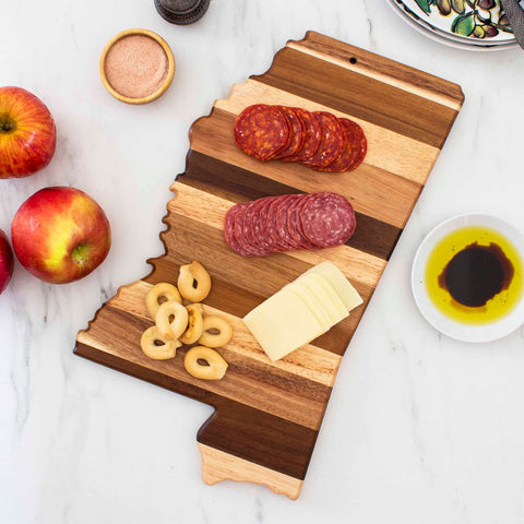 This Rock & Branch® Shiplap Series Mississippi Cutting Board by Totally Bamboo is crafted from rustic wood and shaped like the state of Mississippi. It beautifully displays slices of pepperoni, salami, cheese, and bread rings alongside apples and bowls filled with seasoning, olive oil, and balsamic vinegar.