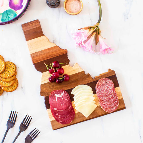 The Rock & Branch® Shiplap Series Idaho Cutting Board by Totally Bamboo features slices of salami, cheese, and cherries on its wooden surface. Nearby are crackers, forks, and a pink flower arranged on a white background. This piece also serves as delightful wall art.