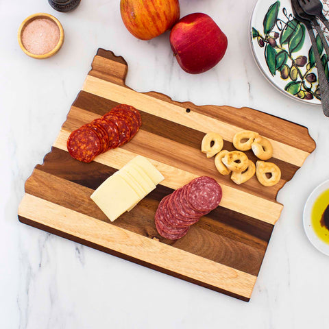 The Rock & Branch® Shiplap Series Oregon Cutting Board by Totally Bamboo is designed in the shape of Oregon with wood planks in varying shades. It stylishly presents sliced pepperoni, cheese, salami, and pretzels. Adjacent to it rests an apple, a small bowl of salt, and plates accompanied by utensils on a marble surface.