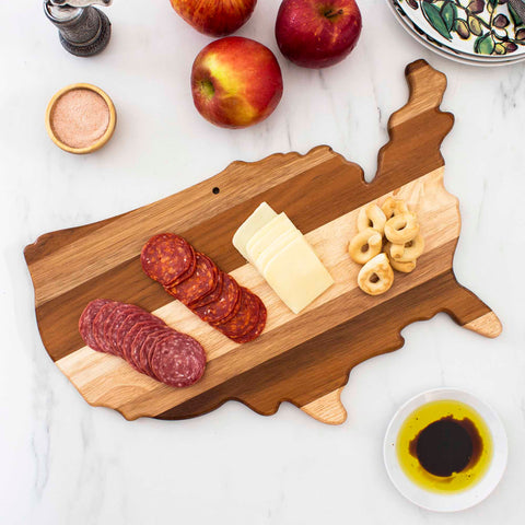 The Rock & Branch® Shiplap Series USA Cutting Board by Totally Bamboo, with its delightful shiplap design, showcases sliced salami, cheese, and round crackers. The display is complemented by apples, a small bowl containing olive oil and vinegar, and a dish featuring a decorative pattern, all elegantly arranged on a marble surface.