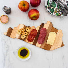 Using the Rock & Branch® Shiplap Series Kentucky Cutting Board from Totally Bamboo, this wooden serving board, designed in the shape of Kentucky, elegantly displays sliced salami, cheese, and round bread snacks. Accompanying it are red apples, a patterned plate with utensils, a pepper grinder, a small bowl containing balsamic vinegar and oil, and another small wooden bowl.