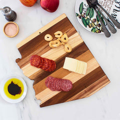 A Rock & Branch® Shiplap Series Arizona Cutting Board from Totally Bamboo showcases sliced salami, pepperoni, cheese, and taralli biscuits. Nearby rests a small dish of olive oil with balsamic vinegar, an apple, a pepper grinder, and a decorative plate with utensils.