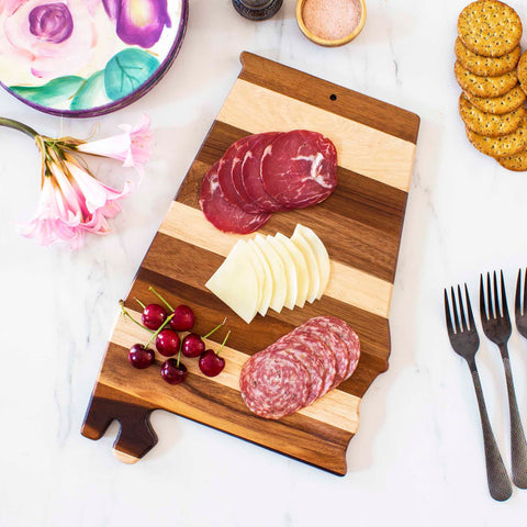 A striped wooden cutting board from the Rock & Branch® Shiplap Series by Totally Bamboo, shaped like Alabama, holds sliced meats, cheese, and cherries. Nearby are pink flowers, a colorful plate, a bowl of salt, crackers, and three forks on a marble surface.