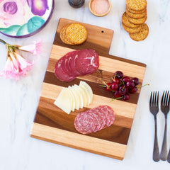 Displayed on the Rock & Branch® Shiplap Series Utah Cutting Board by Totally Bamboo, slices of cured meat, cheese, and cherries create a striking arrangement. Nearby are crackers, a floral plate, a small bowl of salt, two forks, and pink flowers adorning the marble surface.