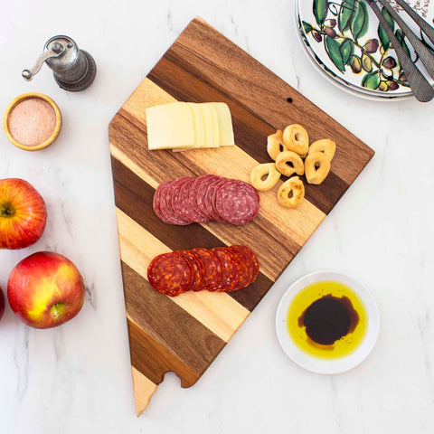 The Rock & Branch® Shiplap Series Nevada Cutting Board by Totally Bamboo displays slices of cheese, salami, pepperoni, and crackers. Nearby, a small bowl of olive oil and balsamic vinegar sits with apples and a set of plates featuring patterned designs on a marble surface.