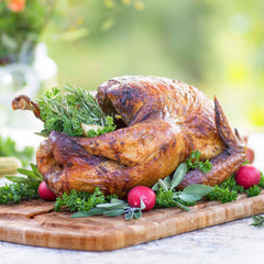 A roasted whole chicken is garnished with fresh herbs such as rosemary, parsley, and sage, alongside vibrant red radishes. It is elegantly presented on the Family Tree Etched Carving Board by Totally Bamboo, measuring 19-1/2" x 15-3/4", set against a blurred green outdoor background.