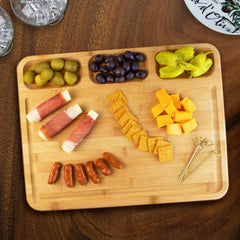 The 3-Well Kitchen Prep Board by Totally Bamboo, measuring 17-1/2" x 13-1/2", presents a charming assortment of snacks perfect for a charcuterie spread: green and black olives, pickled peppers, cheese cubes, crackers, mini sausages, and prosciutto-wrapped cheese sticks. This functional cutting board also includes decorative toothpicks and features a juice groove to capture any drips.