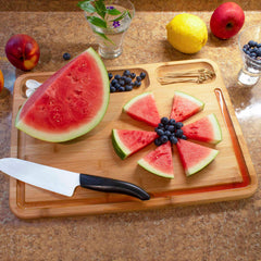 A Totally Bamboo 3-Well Kitchen Prep Board, measuring 17-1/2" x 13-1/2", showcases a vibrant arrangement: sliced watermelon and blueberries placed in a circular pattern with a knife nearby. Whole fruits, including lemons and apples, surround this board with its juice groove feature. A glass filled with small purple flowers completes the display.