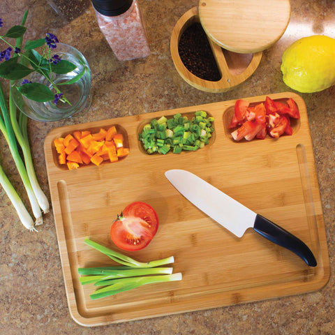 The 3-Well Kitchen Prep Board by Totally Bamboo, measuring 17-1/2" x 13-1/2", elegantly showcases sliced tomatoes, green onions, and orange bell peppers. A nearby knife is accompanied by a salt shaker, spice container, and lemon. A vase with flowers adds charm while the board's juice groove ensures neatness on the bustling counter.