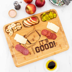 A football-themed "It's Good" 3-Well Kitchen Prep Board by Totally Bamboo, measuring 17-1/2" x 13-1/2", features a football engraving and holds an assortment of sliced meats, cheeses, apple slices, banana chips, and pepperoncini peppers for touchdown-dance snacks. Nearby are a bowl of olive oil, apples, and condiments ready to be served.