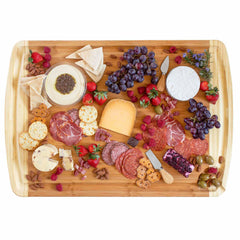 A top view of a Totally Bamboo 600SI Large Cutting Board & Noodle Board, measuring 30" x 20", is elegantly adorned with an assortment of cheeses, meats, grapes, berries, olives, crackers, and nuts. A cheese knife is nestled nearby in the juice groove as several items are artistically arranged on the board.