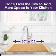 A Totally Bamboo 600SI Large Cutting Board & Noodle Board, measuring 30" x 20", is set over the kitchen sink. The scene includes a backdrop of a white tiled wall, a window revealing lush trees, and an orchid in a pot next to a gleaming silver faucet. Text at the top reads, "Place Over the Sink to Add More Space to Your Kitchen.