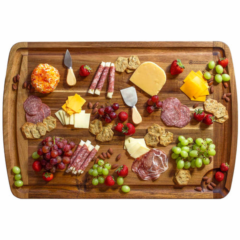 The Rock & Branch® 600SI Cutting Board & Noodle Board, measuring 30" x 20", by Totally Bamboo, elegantly showcases an array of cheese, cured meats, grapes, strawberries, crackers, and almonds. Cheese knives add the perfect touch to the presentation on the charcuterie board, emphasizing the diverse textures and vibrant colors displayed.