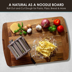 The Rock & Branch® 600SI Cutting Board & Noodle Board, measuring 30" x 20", from Totally Bamboo, is showcased as a noodle board featuring a pasta machine, a nest of fresh pasta, cheese block, garlic, parsley bunch, red onions, and tomatoes. The text above states: "A Natural as a Noodle Board" with an emphasis on rolling and cutting dough.