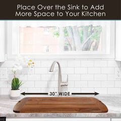 The Rock & Branch® 600SI Cutting Board & Noodle Board by Totally Bamboo, measuring 30 inches by 20 inches, extends across a kitchen sink and serves as both a cutting surface and charcuterie board. It elegantly rests on a marble countertop framed by a subway tile backsplash. A potted orchid and plant enhance the scene, with the window providing a view of the trees outside.