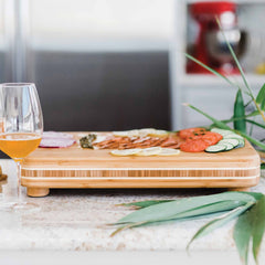 The "Big Easy" Chopping Block by Totally Bamboo, measuring 19" x 13-1/2", displays an assortment of sliced vegetables, salami, and herbs atop a kitchen counter. A glass filled with an amber-colored drink enhances the ambiance. In the background, a blurred kitchen shows off a mixer, while in the foreground are potted plant leaves.