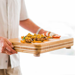 A person holding a "Big Easy" Chopping Block, measuring 19" x 13-1/2", from Totally Bamboo, adorned with assorted food items like sliced tomatoes, vegetables, and other garnishes. The individual is wearing a white shirt that highlights the vibrant and softly blurred background, suggesting an artful ease in food preparation.