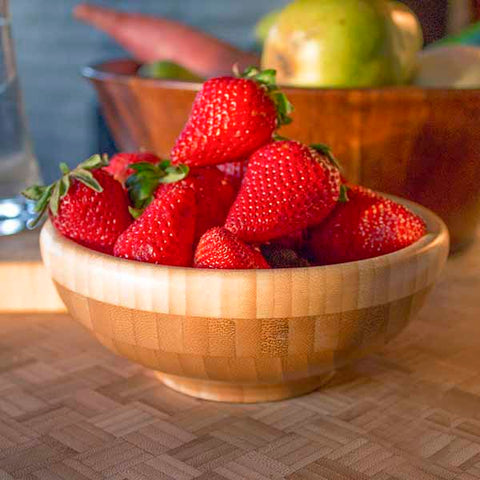 A timeless 6" Classic Bowl by Totally Bamboo, crafted from durable Moso bamboo, holds vibrant red strawberries on a wooden table. The bowl beautifully contrasts with the fruit's rich color. In the background, another bowl fades into a blur, featuring an assortment of other fruits. The strawberries proudly display their green leaves.