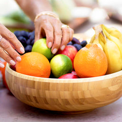 Hands arranging a selection of fresh fruit, such as oranges, limes, bananas, and berries, in the Totally Bamboo 12" Classic Bowl that serves as a vibrant centerpiece.