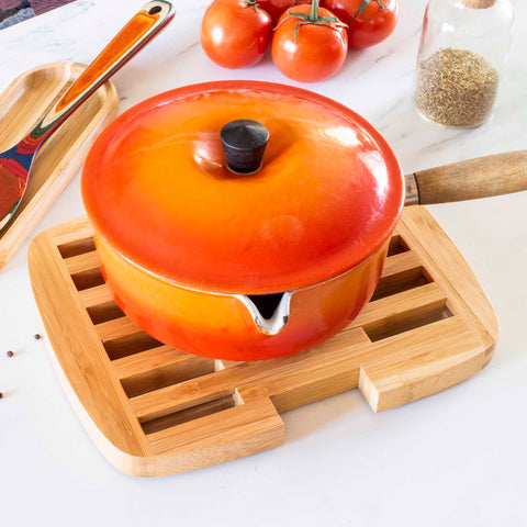 A round orange pot with a black handle is placed on the Totally Bamboo Expandable Trivet for Hot Plates, which expands to 11-1/2" wide and provides countertop protection. Fresh tomatoes, a wooden spoon, and a small jar of herbs decorate the marble countertop nearby.