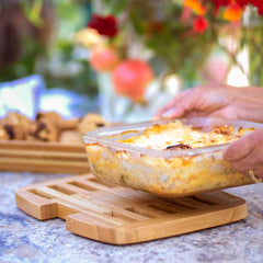 A person places a glass dish with baked casserole on the Totally Bamboo Expandable Trivet for Hot Plates, which expands up to 11-1/2" wide. The background features blurred flowers and cookies on a wooden tray, protecting the countertops and enhancing the suggestion of a warm and inviting outdoor meal setting.