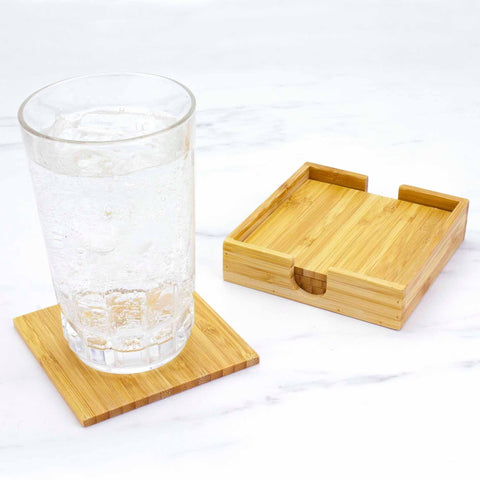 A glass of sparkling water with ice rests on a wooden coaster crafted from Moso bamboo. Nearby, a stylish Bamboo Coaster Set holder from the Totally Bamboo 4-Piece Bamboo Coaster Set with Case enhances the white marble surface.