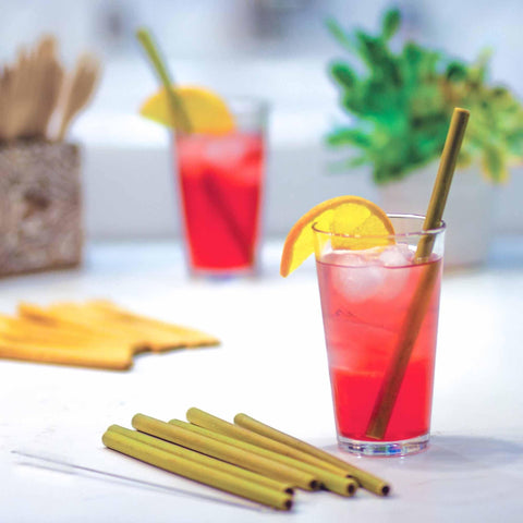 On a white table, a vibrant red drink in a glass, adorned with an orange slice and cooled by ice, is surrounded by the Totally Bamboo Reusable Bamboo Drinking Straws from an 8 pack. A second glass and a potted plant are softly out of focus in the background, highlighting the chic sustainability of these eco-friendly bamboo straws as renewable resources.