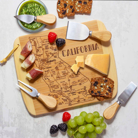 A Totally Bamboo A Slice of Life California Cutting Board, measuring 11" x 8-3/4", is perfect for presenting an elegant snack display featuring cheese, prosciutto-wrapped bites, nuts, and berries. It's beautifully complemented by surrounding cheese knives, crackers, and a small bowl of pesto for a delightful State Outline-inspired feast.