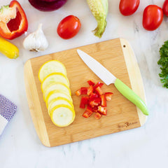 An 11" x 8-3/4" A Slice of Life New Jersey Cutting Board by Totally Bamboo, along with a green-handled knife, displays sliced yellow squash and chopped red bell pepper. Nearby on the marble countertop are tomatoes, garlic, corn, lettuce, a red onion, and an uncut yellow squash.