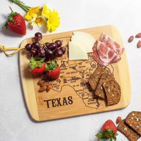 On the Totally Bamboo "A Slice of Life Texas" Cutting Board, measuring 11" x 8-3/4", sits a beautifully arranged charcuterie board featuring sliced cheese, cured meat, grapes, strawberries, almonds, and crackers. Nearby, bright yellow flowers add an elegant and stylish touch to the display.